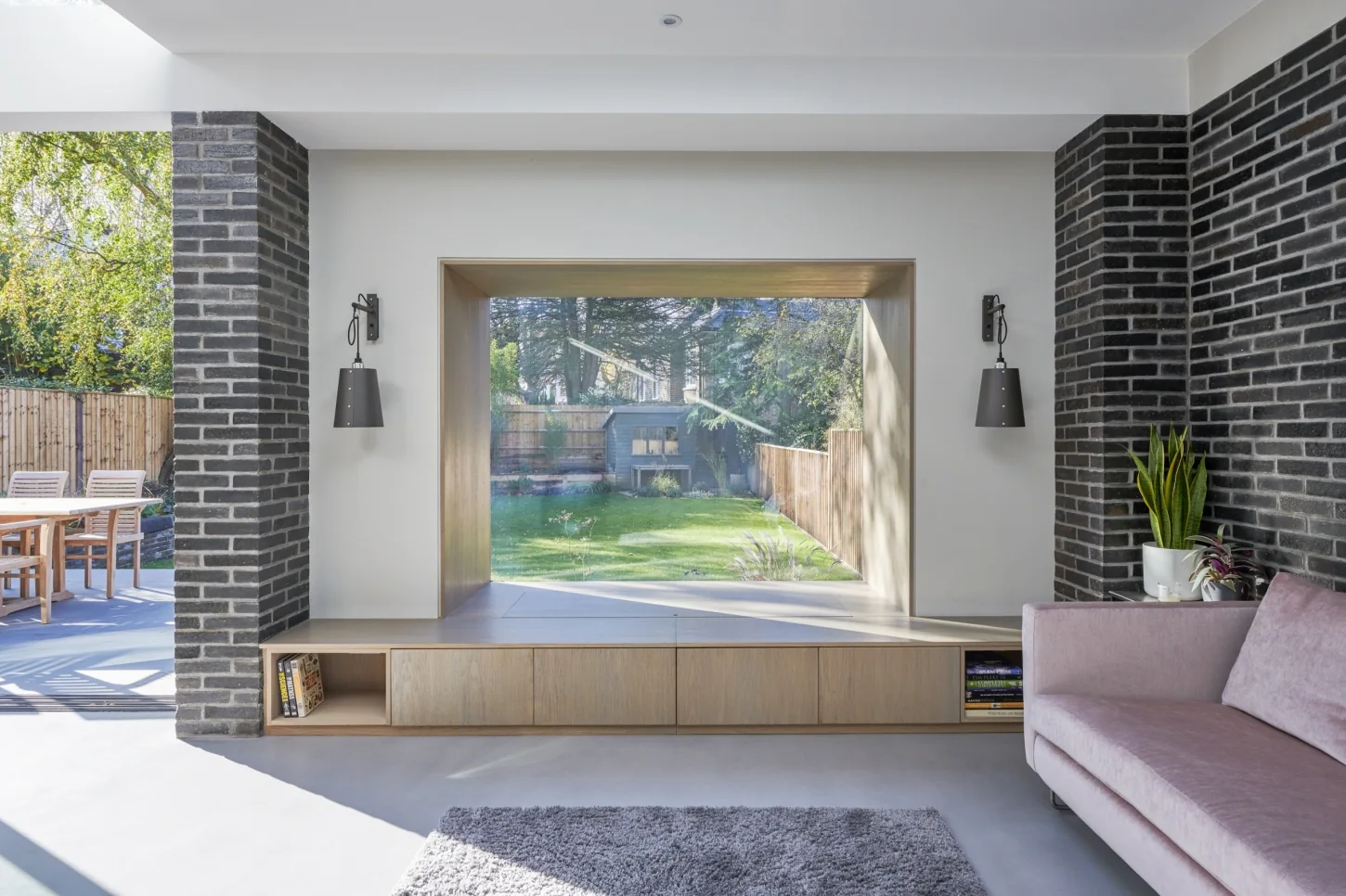 Interior view through a Maxlight Oriel Bay Window — an oak-lined projecting bay with frameless glass-to-glass corners framing a garden, set between brick walls above a low timber bench.