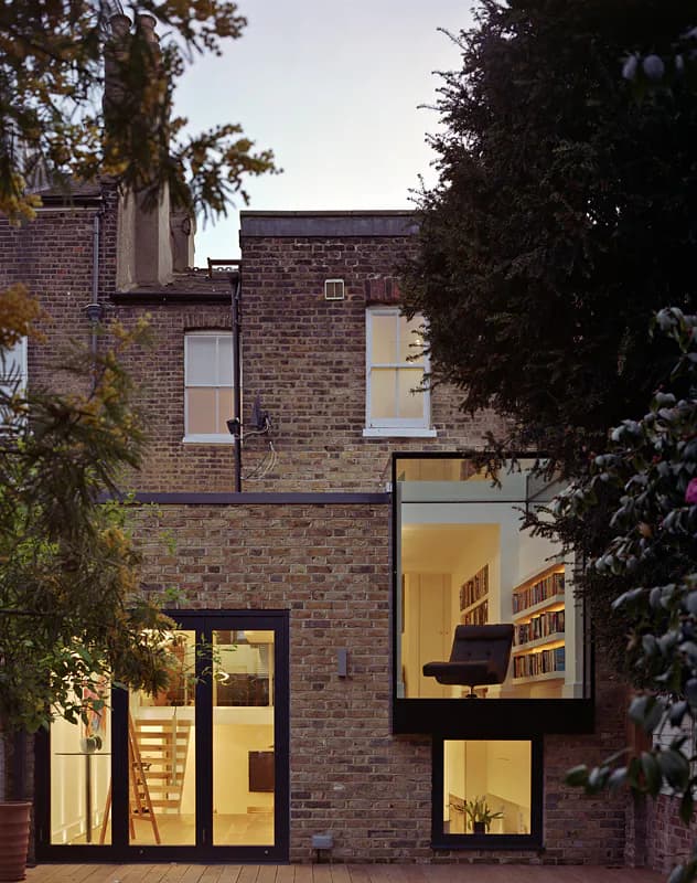 Projecting glass reading room glowing on the rear of a brick house at dusk